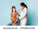 Coronavirus Vaccination And Pregnancy. Nurse Vaccinating Pregnant Female Patient Against Covid-19 Sticking Bandage After Vaccine Injection In Arm Over Blue Studio Background, Wearing Face Masks
