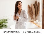 Haircare Concept. Joyful Pretty Woman In Bathrobe Touching Long Brunette Hair Standing In Modern Bathroom At Home In The Morning. Lady Enjoying Beauty Routine Caring For Herself