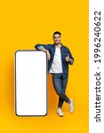 Small photo of Happy arab guy leaning on huge smartphone with empty screen and showing thumb up, mockup, yellow studio background, full length shot, copy space, mobile application, advertisement concept