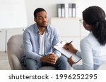 Unhappy young black man having session with professional psychologist at mental health clinic. Psychotherapist taking notes during conversation with depressed male patient