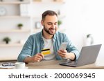 Happy Caucasian man buying things online, using smartphone, laptop and credit card, enjoying shopping in internet. Positive young guy purchasing goods on web, making remote payment