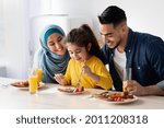 Tasty Breakfast. Happy Islamic Parents And Little Daughter Eating Together In Kitchen, Adorable Female Child Using Fork While Tasting Food, Happy Mom And Dad Looking At Their Kid, Closeup Shot