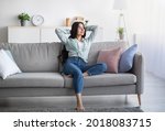 Small photo of Full length of young Indian lady relaxing on lazy weekend at home, sitting on couch with hands behind her head, copy space. Young woman having day off, chilling on comfy sofa indoors