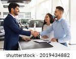 Smiling middle-eastern young man and woman buying car at auto salon, handsome arab guy shaking sales assistant hand, making successful deal, happy family got brand new luxury car, side view