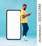 Small photo of Full length of young Afro guy using cellphone while leaning on giant mobile phone with empty white screen, blue studio background. Mockup for app or website, space for advertisement
