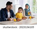 Happy arab family of three eating tasty lunch together at home, cheerful islamic parents and little daughter sitting at table in kitchen, enjoying healthy delicious food and drinking orange juice
