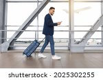 Small photo of Handsome arab guy using smartphone while walking with suitcase at airport terminal, young middle eastern man browsing mobile internet on cellphone while going to flight boarding, side view
