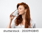 Healthy Hydration. Red-Haired Millennial Woman Drinking Water From Glass Standing Over Gray Studio Background, Wearing Casual T-Shirt. Side View Shot. Stay Hydrated Concept