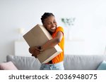 Cheerful Afro woman hugging carton parcel, receiving long awaited delivery, getting online order indoors. Satisfied female customer empracing her internet purchase in cardboard package