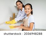 Young asian mother teaching her daughter housekeeping, cleaning kitchen together and smiling at camera, selective focus. Cute girl wiping table with cloth, enjoying helping with household chores