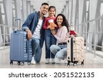 Small photo of Happy Middle Eastern Family Of Three Posing In Airport Terminal, Cheerful Arab Parents And Their Cute Little Daughter Holding Suitcases, Passports And Tickets And Smiling At Camera, Ready For Travel