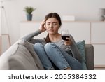 Small photo of Sad indifferent caucasian young lady sits on sofa with glass of wine and suffering from headache in home interior. Alcohol addiction, alcoholism, hangover, loneliness and depression, mental health