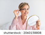 Natural Makeup Concept. Attractive Lady Applying Mascara On Eyelashes Enjoying Morning Beauty Routine Holding Mirror And Looking At Camera. Female Cosmetics Product, Free Copy Space