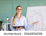 Small photo of Happy French teacher explaining foreign language rules near blackboard indoors and smiling at camera. Confident young female tutor teaching linguistic online course