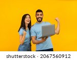 Small photo of Overjoyed arab couple holding laptop and celebrating success with clenched fists, lucky winners posing with PC computer over yellow studio background, copy space