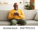 Small photo of Senior Black Man Using Smartphone Texting Networking Online And Scrolling News In Social Media Application Sitting On Couch At Home. Mature Male Reading Message On Phone. New Mobile App Concept