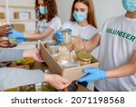Small photo of Young volunteers in masks packing food in cans, jars and packages into carton boxes as donation for poor people. Group of millennials working in charitable foundation