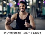 Small photo of Fitness Nutrition. Happy Muscular Arab Man Holding Sport Shaker And Protein Bar While Relaxing After Training At Gym, Smiling Middle Eastern Male Athlete Having Break In Workout, Enjoying Snacks