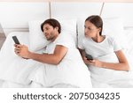 Small photo of Disgruntled distrustful european young wife looks at husband phone, guy chatting in social networks in bed in white bedroom interior, top view. Suspicion of treason, betrayal, relationship problems