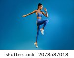 Female Athlete Jumping Exercising During Training Over Blue Studio Background, Looking Aside. Fitness Workout And Sport Motivation Concept. Full Length, Side View