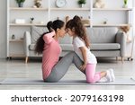 Small photo of Loving middle eastern mother and daughter in sportswear working on abs together, exciting little girl holding her mom legs while she doing crunches, touching each other with foreheads, home interior