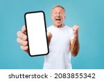 Small photo of Big Luck And Win. Excited Mature Male Holding And Showing Big Cell Phone With Empty White Screen In Hand For Mock Up, Shaking Clenched Fist, Cheerful Man Celebrating Win, Standing At Blue Studio