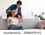 Smiling millennial european guy closes eyes to woman, gives present in box to surprise smiling wife in room interior, empty space. Party at home, birthday gift, anniversary, Valentines day and holiday