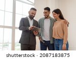 Small photo of Millennial Couple Buying New Apartment, Standing In Empty Room, Professional Real Estate Agent In Suit Showing Digital Tablet With Build Project, Discussing House Plan, Selling Flat To Happy Family