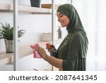 Small photo of Hygiene And Cleanliness. Portrait of happy muslim lady in hijab cleaning bookshelf surface in living room, bedroom or office, wiping dust holding spray bottle and using microfiber cloth