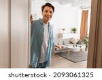Portrait of cheerful man welcoming inviting visitor to enter his home, happy young guy standing in doorway of modern apartment, millennial male holding door looking out showing living room with hand