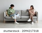 Handsome young Asian guy and his girlfriend using modern gadgets while sitting on couch at home, copy space. Full length of millennial couple communicating online via laptop and tablet pc