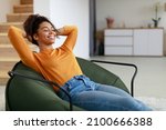 Rest Concept. Happy black lady sitting on comfortable bean bag at home in living room. Cheerful casual woman with closed eyes relaxing on sofa, leaning back, enjoy weekend free time or break from work