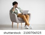 Handsome young indian guy independent contractor working online from home, sitting in comfy armchair, using laptop and having phone conversation with business partner, copy space, full length shot