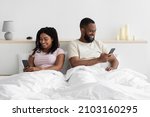 Small photo of Glad young black female ignores her husband sitting on bed and look at phone in bedroom interior, copy space. Treason, addiction on social networks and technology, personal information and new normal
