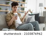 Small photo of Confused asian man looking at phone screen, sitting on couch with computer on lap in living room, upset frustrated male reading bad news on message, having problem with device or Internet connection