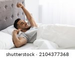 Lazy Morning. Happy Pleased Middle Eastern Man Stretching In Bed After Nice Sleep, Cheerful Young Arab Guy Waking Up With Good Mood, Relaxing In Light Bedroom, Enjoying Weekend Pastime, Copy Space