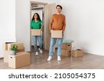 Small photo of Family Housing Concept. Happy couple walking in new apartment and looking around, smiling young guy and lady holding cardboard boxes standing in modern home. Real estate dwelling, mortgage
