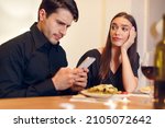 Small photo of Bad Date. Unhappy Sad Beautiful Woman Is Getting Bored Sitting On Date Event In Restaurant While Her Boyfriend Using Mobile Phone And Chatting, Ignoring His Girlfriend. Relationship Problem Concept