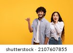Small photo of Omg, check this. Emotional indian guy pointing finger aside at free space over yellow background, surprised lady looking at camera, panorama. Couple demonstrating place for your text