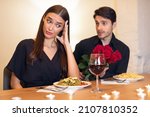 Small photo of Bad Date. Young Couple Having Unsuccessful Meeting In Restaurant, Funny Disappointed Shoked Woman Feeling Embarrassment Covering Face, Man Giving Her Bouquet Of Red Roses