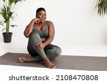 Excited overweight young black woman in tight sportswear sitting on yoga mat and smiling at camera, practicing yoga at home in the morning, panorama with copy space. Healthy lifestyle concept