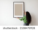 Happy Woman Hanging Empty Poster In Frame On White Wall Standing Indoors. Modern Art, Domestic Interior And Home Decor Pictures Concept. Mockup For Advertisement