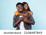 Small photo of Couple In Love. Portrait of joyful black woman hugging her boyfriend from behind, standing together isolated over blue studio background. Casual guy and lady smiling, posing and looking at camera