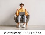 Small photo of Millennial Asian man having creative idea, gesturing eureka with both hands, pointing up, offering space for design, sitting in armchair with laptop pc against white studio wall, copy space