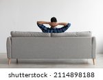 Rear view of young guy sitting on comfortable couch at home in living room, looking at wall. Casual man relaxing on sofa, leaning back holding hands behind head, enjoying weekend free time or break