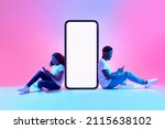 Small photo of Cool mobile app. African American couple sitting near big smartphone with mockup, using modern gadgets, advertising new application or website, promoting your product or service in neon light