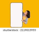 Small photo of Great Offer. Portrait of excited asian woman peeking out big giant vertical cell phone with white blank screen and looking at device display. Gadget with empty free space mock up, yellow orange wall