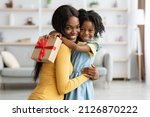 Small photo of Mother's Day Concept. Adorable Female Child Holding Gift And Embracing Mom, Portrait Of Happy African American Mommy And Her Cute Little Daugher Bonding Together At Home, Copy Space