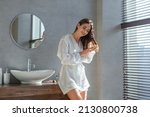 Small photo of Beautiful Young Female In White Silk Robe Brushing Her Hair With Comb While Standing In Modern Loft Bathroom Interior, Happy Millennial Woman Making Daily Beauty Routine At Home, Copy Space