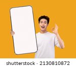 Small photo of Online Offer. Cheerful asian guy demonstrating big smartphone with blank white screen and showing ok gesture at camera, millennial man recommending new mobile app or website, creative collage, mockup